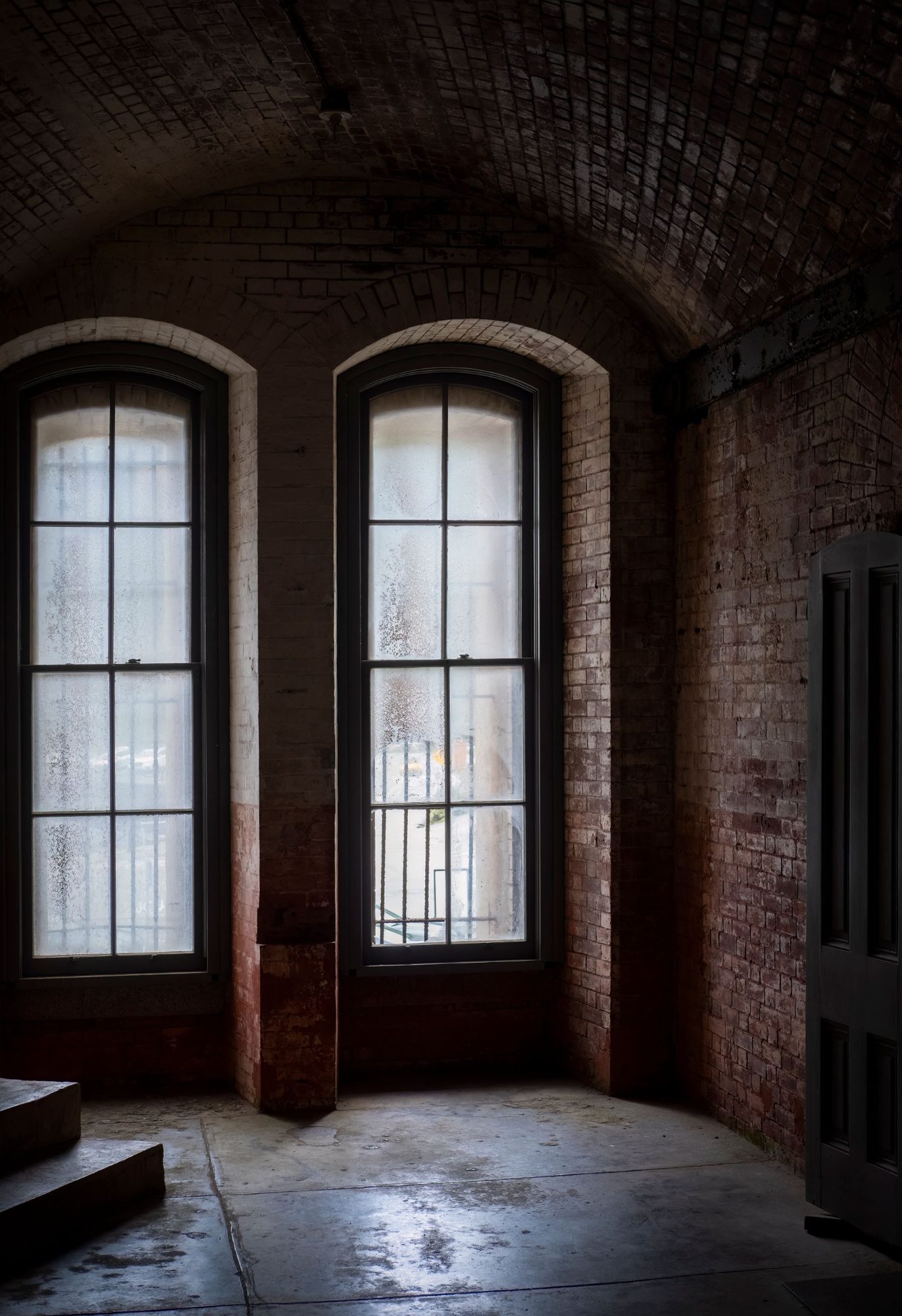 An unused brick-vaulted room lit by two tall arched sash windows